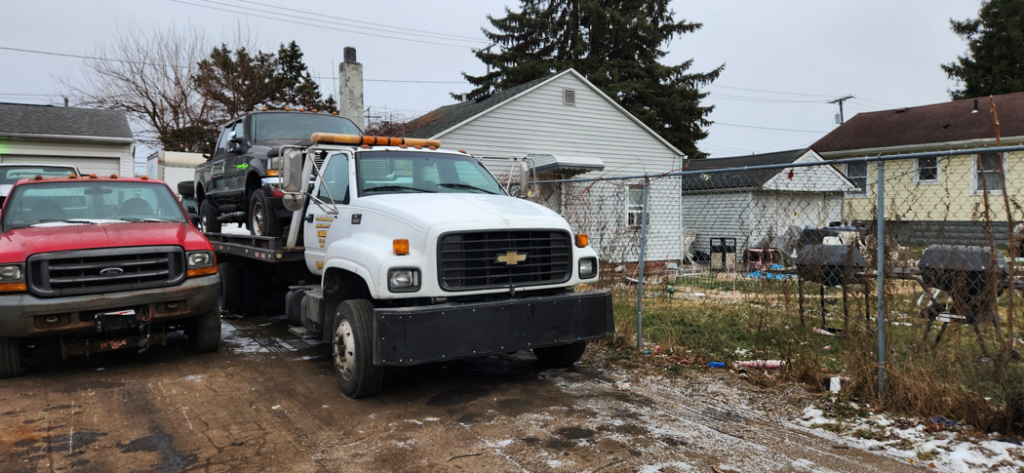 A white flatbed tow truck transporting a dark pickup truck for Boyz Automotive & Towing in Toledo, OH.