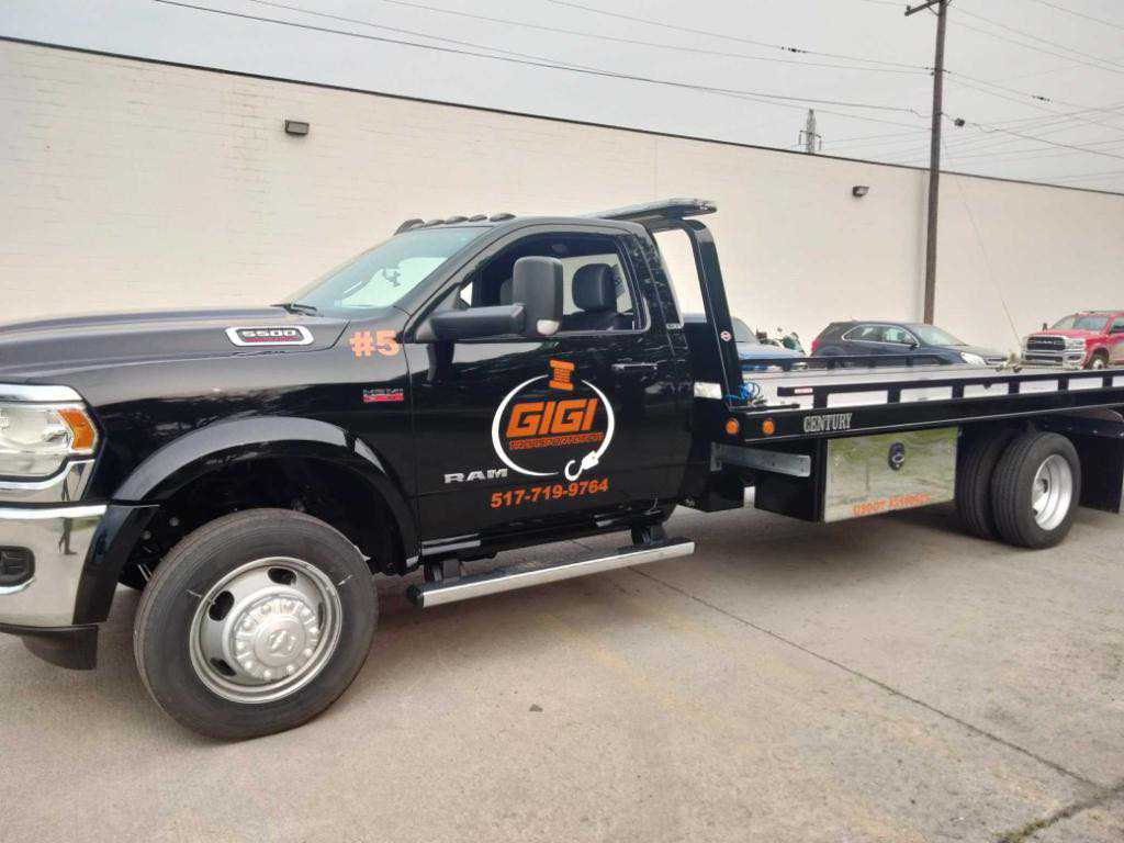 A white flatbed tow truck from Gigi Transportation transporting a dark grey pickup truck in Grand Rapids, MI.