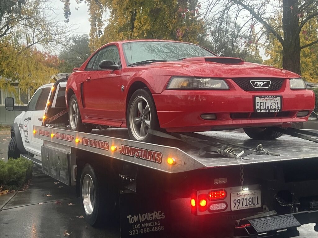A white flatbed tow truck from Kennedy Towing & Transport in Anderson, CA, transporting a red Ford Mustang on a rainy day.
