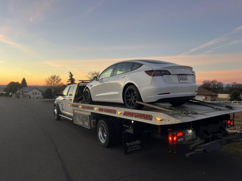 A white flatbed tow truck from Kennedy Towing & Transport in Anderson, CA, transporting a white Tesla Model 3 at sunset.