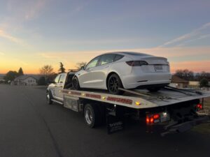 A white flatbed tow truck from Kennedy Towing & Transport in Anderson, CA, transporting a white Tesla Model 3 at sunset.