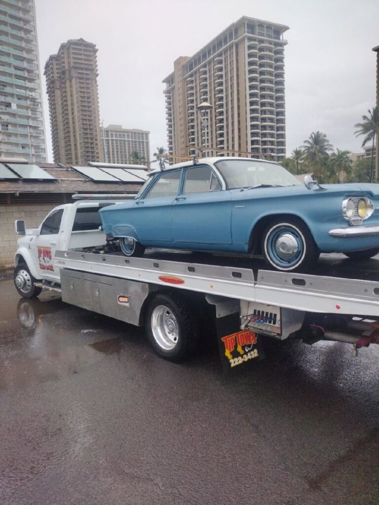 A white Tip Tows flatbed tow truck transporting a blue vintage car in Honolulu, HI.