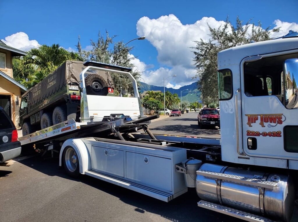 A white Tip Tows flatbed tow truck transporting a large military-style vehicle in Honolulu, HI.