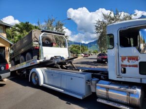A white Tip Tows flatbed tow truck transporting a large military-style vehicle in Honolulu, HI.