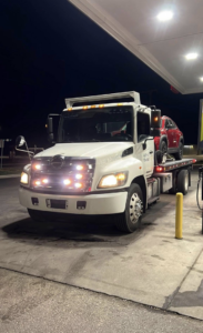 A white flatbed tow truck from BUVO towing services with a red SUV on its bed at a gas station at night in Fort Worth, TX.