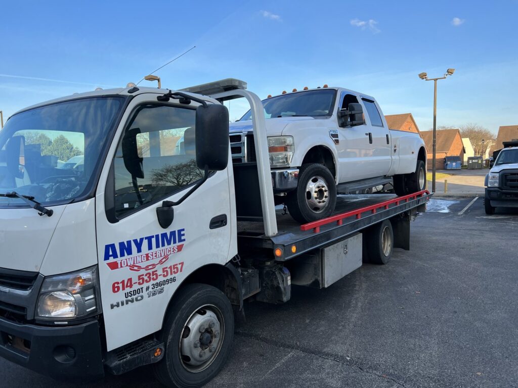A white Ford F-350 dually pickup truck on a flatbed tow truck with the Anytime towing services logo in Columbus, OH.