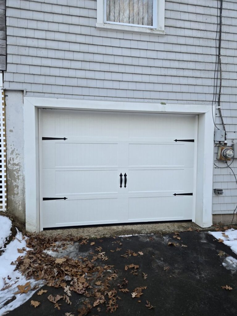 A newly installed white garage door with decorative hardware on a gray-sided house by Spring King Garage Doors in Middletown, CT