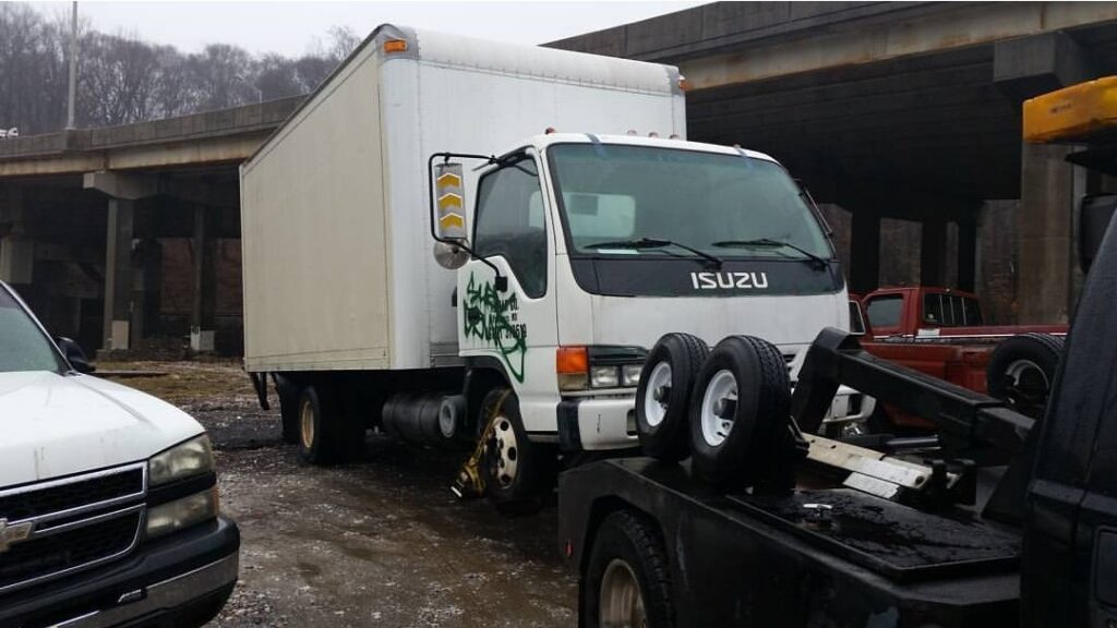A white Isuzu box truck being towed by Whitelock Towing in Baltimore, MD, using a tow dolly.