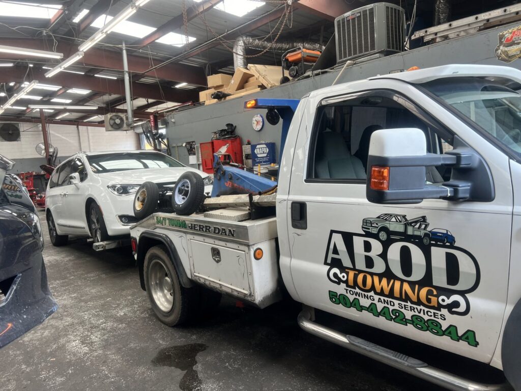 A white minivan being towed by a wheel lift tow truck from ABOD Towing LLC in New Orleans, LA, inside a repair shop.
