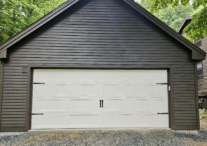 A white panel garage door with decorative black hardware installed on a dark brown house by Champion Overhead Door in Brattleboro, VT.