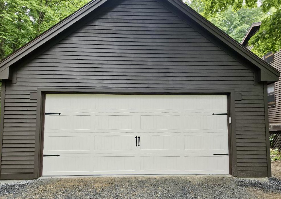 A white panel garage door with decorative black hardware installed on a dark brown house by Champion Overhead Door in Brattleboro, VT.