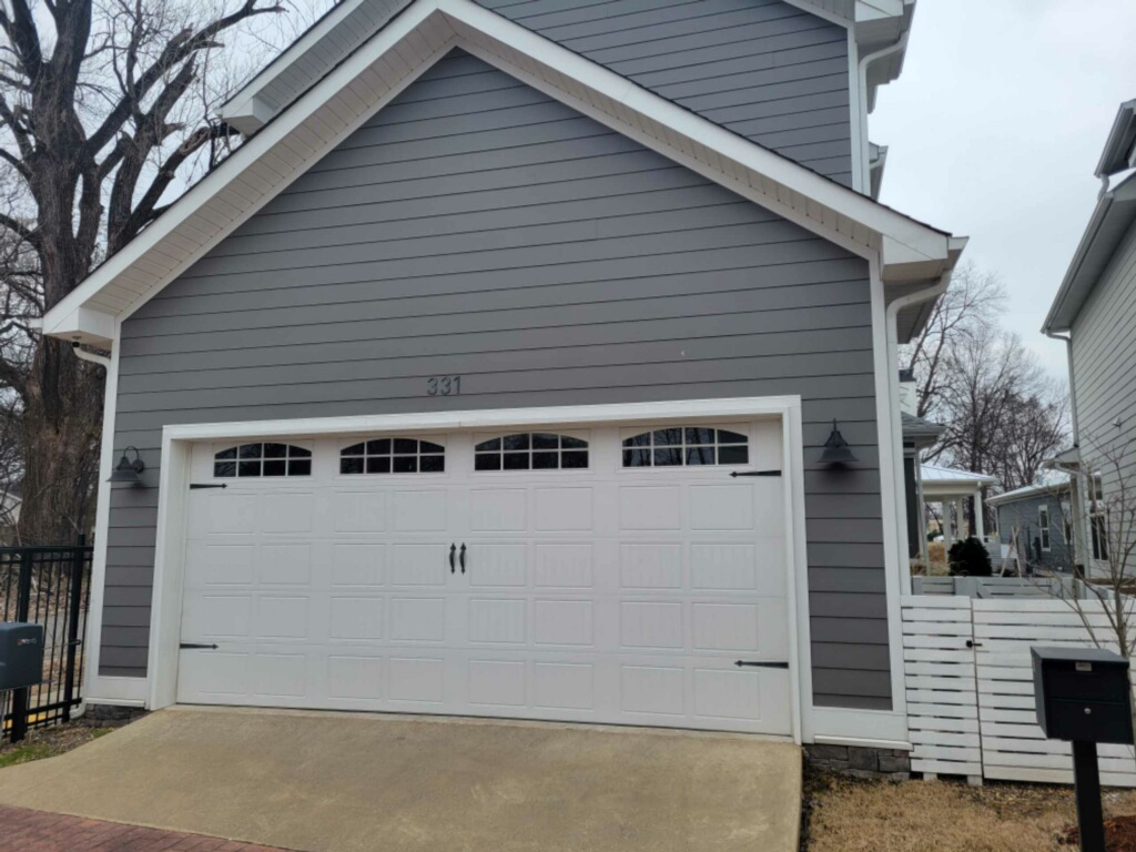A newly installed white paneled garage door with decorative windows on a grey house by Garage Door Sales & Repair in Conway, AR.