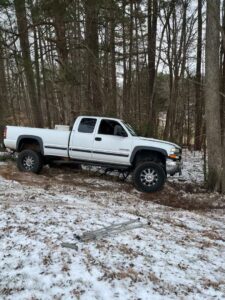 A white pickup truck stuck in a snowy, wooded area, indicating a recovery service by Bullcity Towing & Services LLC in Durham, NC.