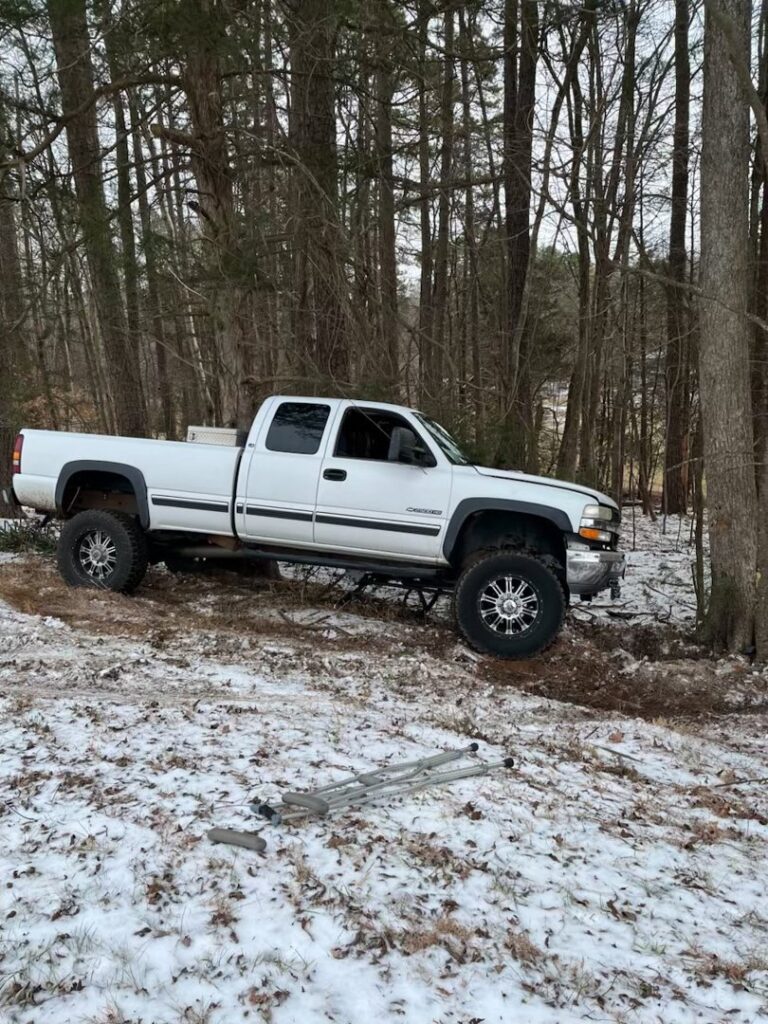 A white pickup truck stuck in a snowy, wooded area, indicating a recovery service by Bullcity Towing & Services LLC in Durham, NC.