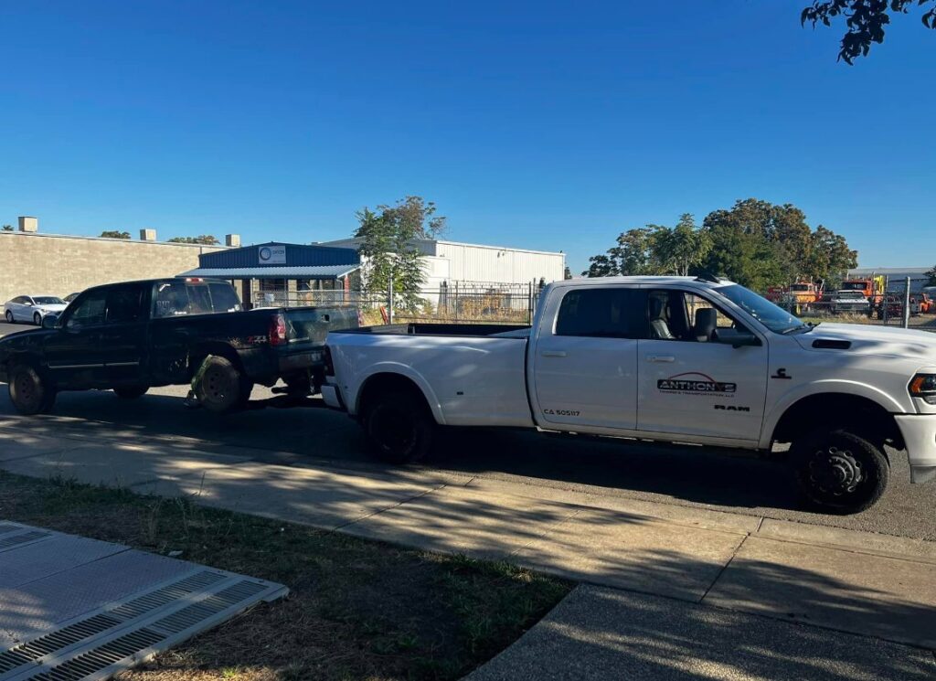 A white Anthony's Towing & Transportation pickup truck actively towing a dark pickup truck in Yuba City, CA.