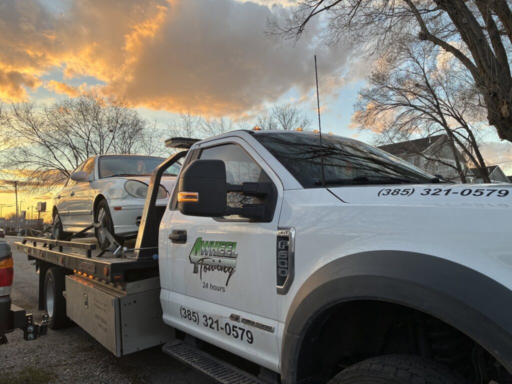 A white sedan being transported on a flatbed tow truck by 4 Wheel Towing at sunset in South Salt Lake, UT.