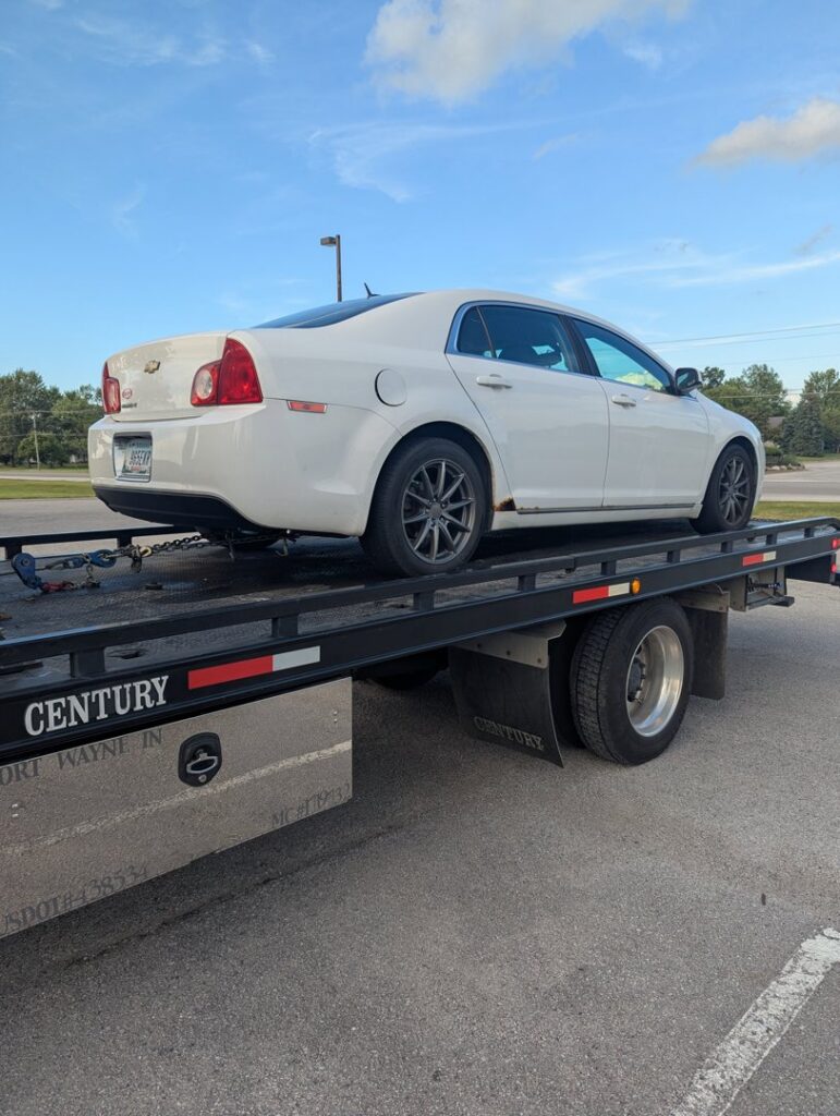 A white sedan being transported on a flatbed tow truck by First Hand Towing & Recovery in Fort Wayne, IN.