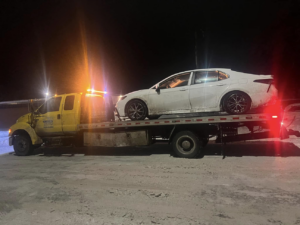 A white sedan loaded onto a flatbed tow truck by Geely Towing at night in Fargo, ND.