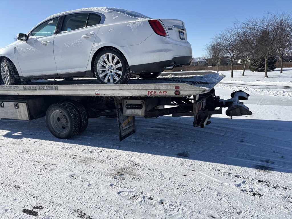 A white sedan being transported on a flatbed tow truck in a snowy environment by Towing Recovery Rebuilding Assistance Services in Naperville, IL