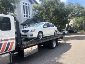 A white Nissan Sentra sedan being transported on a TP Towing flatbed truck in a residential area of Tampa, FL.
