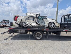A white severely damaged car being recovered and transported on a flatbed tow truck by First Hand Towing & Recovery in Fort Wayne, IN.