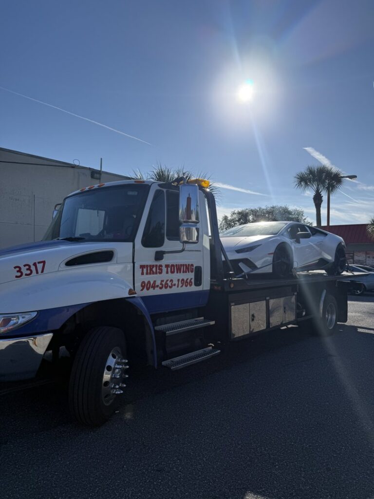A white sports car being transported on a flatbed tow truck by Tiki's Towing LLC in Jacksonville, FL.
