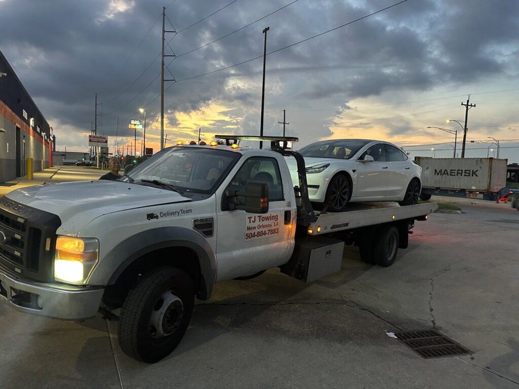 TJ Towing LLC flatbed truck towing a white Tesla Model 3 at dusk in New Orleans, LA.