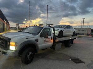 TJ Towing LLC flatbed truck towing a white Tesla Model 3 at dusk in New Orleans, LA.