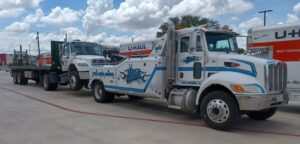 A white tow truck from Alanis Wrecker Service towing a flatbed truck with a U-Haul trailer in San Antonio, TX.