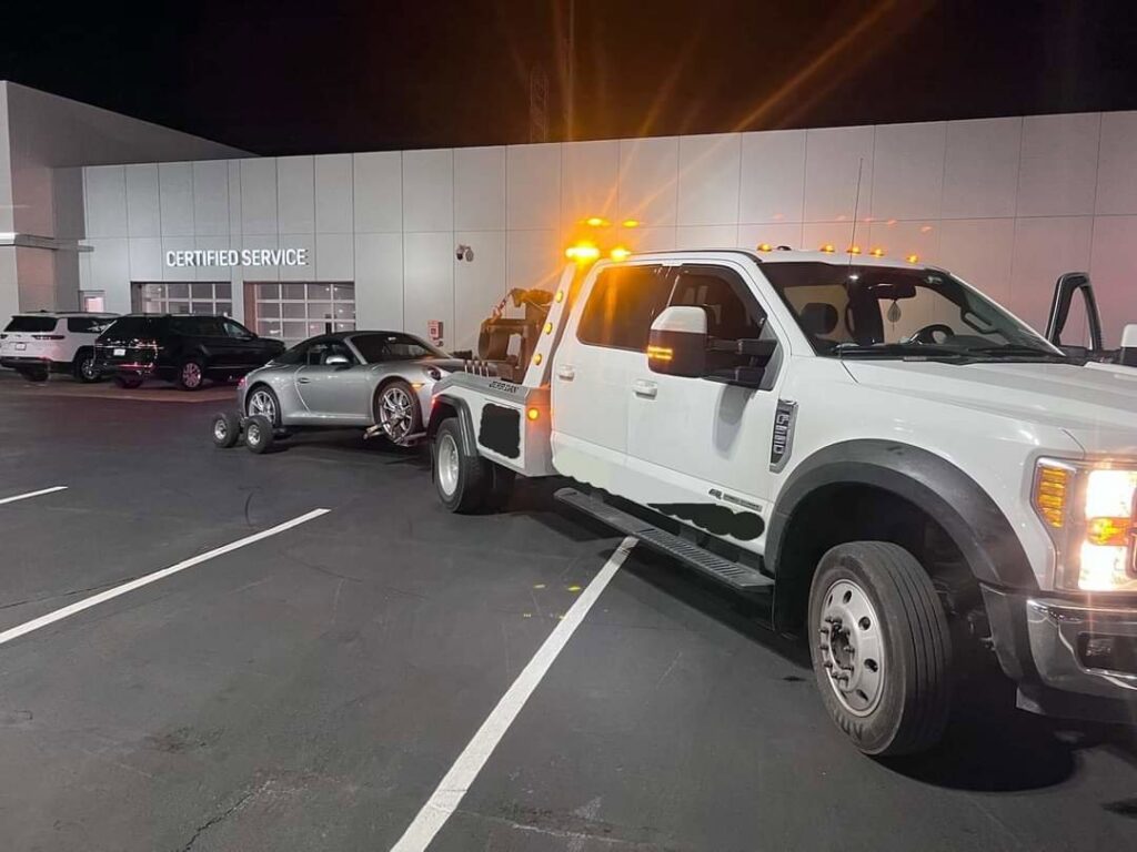 A white tow truck towing a silver sports car with dolly wheels at night by Towing Recovery Rebuilding Assistance Services in Naperville, IL