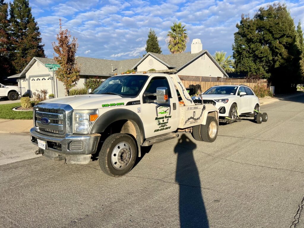 A white tow truck from Kennedy Towing & Transport in Anderson, CA, towing a white SUV on a dolly in a residential area.
