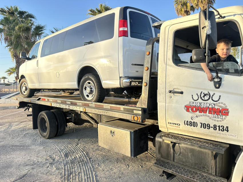 A white van being transported on a flatbed tow truck by Trejo's Towing LLC in Phoenix, AZ, with a child in the tow truck's cab.