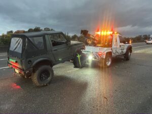 A white wheel-lift tow truck from Kennedy Towing & Transport in Anderson, CA, towing a green off-road vehicle on a highway at dusk.