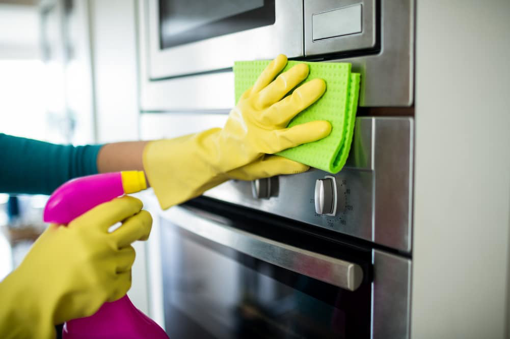 A person wearing yellow gloves wipes a stainless steel oven with a green cloth, showcasing house cleaning by The Cathartic Couple in Cheyenne, WY.