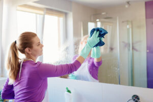A woman wearing gloves and an apron, cleaning a bathroom mirror for Clean Deeds in Olympia, WA.