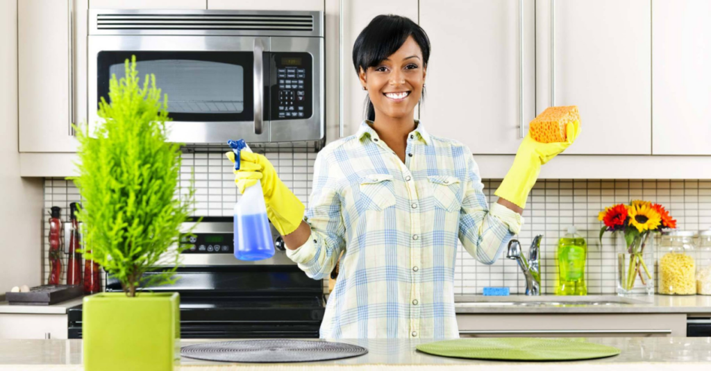A smiling woman wearing yellow gloves, holding a spray bottle and sponge, ready to clean a kitchen for Page not active in St. Charles, IL.