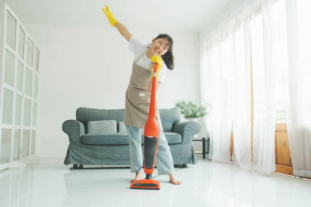 A smiling woman in an apron and gloves vacuuming a living room floor, representing house cleaning services by Cascade Fresh Cleaning Services in Grand Rapids, MI.