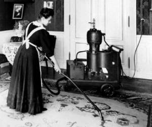 A woman in a maid's uniform vacuuming a rug with an antique vacuum cleaner for Carnation Services LLC in West Allis, WI.