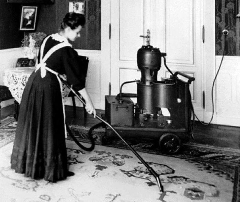 A woman in a maid's uniform vacuuming a rug with an antique vacuum cleaner for Carnation Services LLC in West Allis, WI.