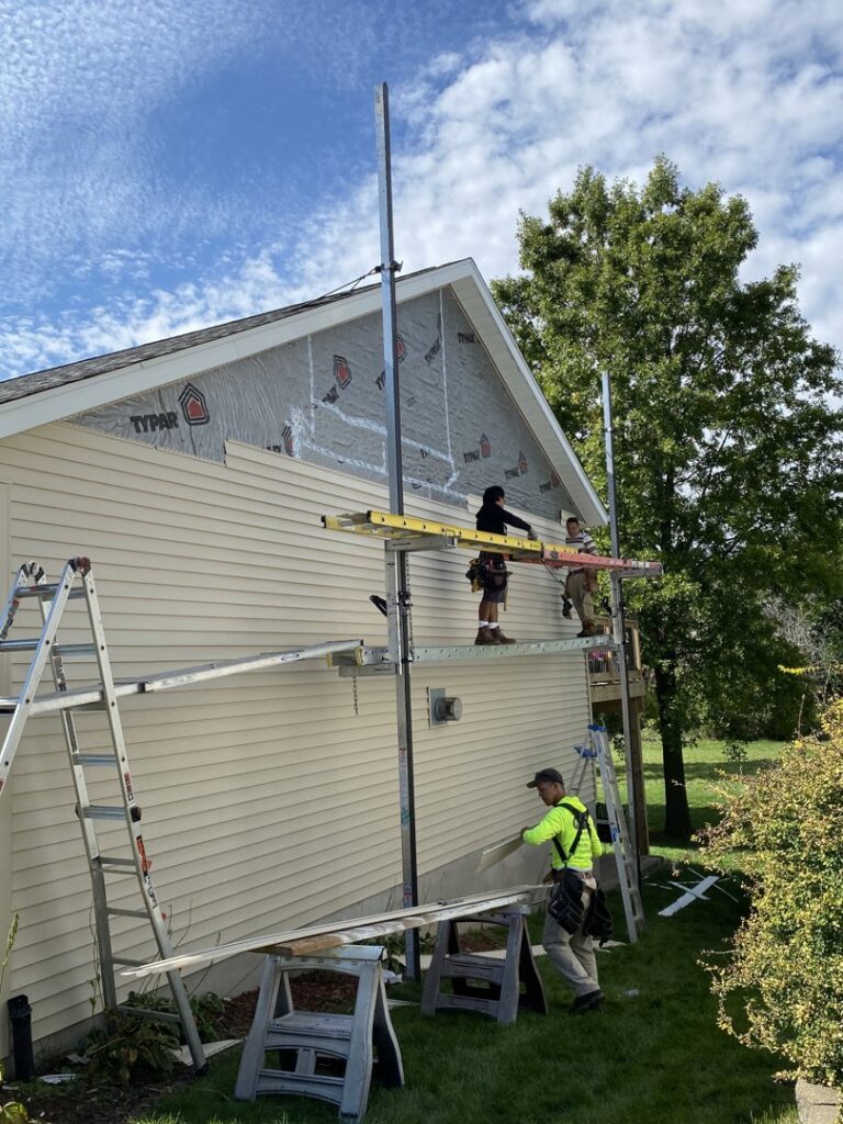 A worker on a ladder installing red siding on a house with a bay window, by DJ Paz All Siding & Roofing Construction in Iowa City, IA