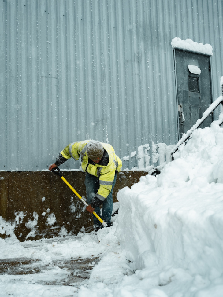 A worker in a high-visibility jacket shoveling snow by hand next to a building for Elcor Construction in Rochester, MN.