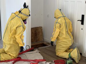 Two workers in yellow hazmat suits inspecting an opening in the floor during a water damage restoration job by Restoration 1 of OKC in Yukon, OK.