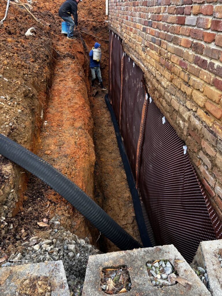 Workers installing foundation waterproofing membrane and drainage pipe next to a brick house by Stay Dry Waterproofing LLC in Marysville, OH.