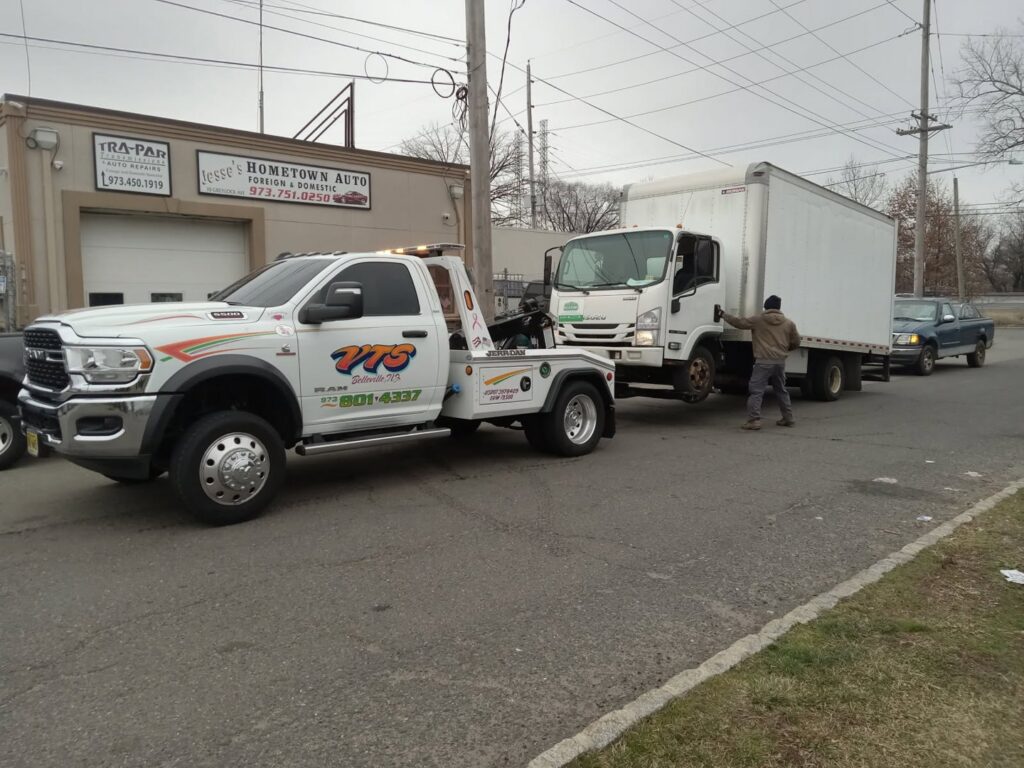 A Velez Towing wrecker truck actively towing a white box truck with a trailer on a street in Newark, NJ.