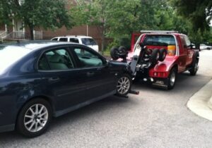 A wrecker tow truck from Raleigh Towing & Recovery towing a dark sedan with its front wheels lifted in Raleigh, NC.