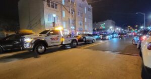 A Velez Towing wrecker truck towing a silver car on a city street at night in Newark, NJ.
