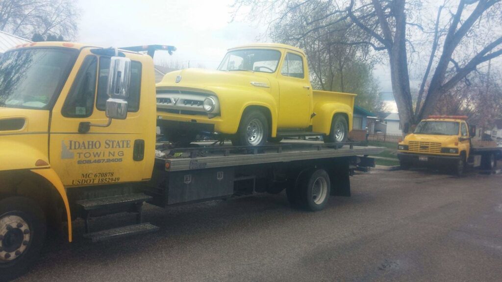 A yellow classic pickup truck being transported on a flatbed tow truck by Idaho State Towing and Recovery in Caldwell, ID.