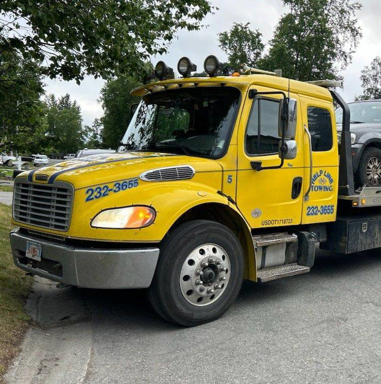 A yellow flatbed tow truck from Help Line Towing transporting a car on a street in Wasilla, AK.