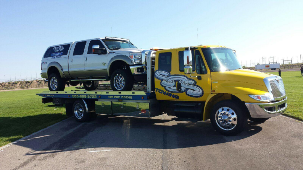 A yellow flatbed tow truck from SOS Towing carrying a silver pickup truck in Albuquerque, NM.