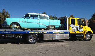A yellow flatbed tow truck from SOS Towing carrying a light blue vintage car in Albuquerque, NM.